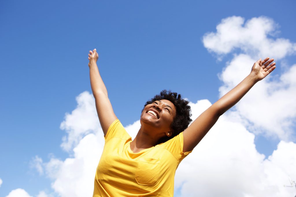 Woman in bright yellow shirt smiling joyfully, throwing her hands toward blue sky on a sunny day