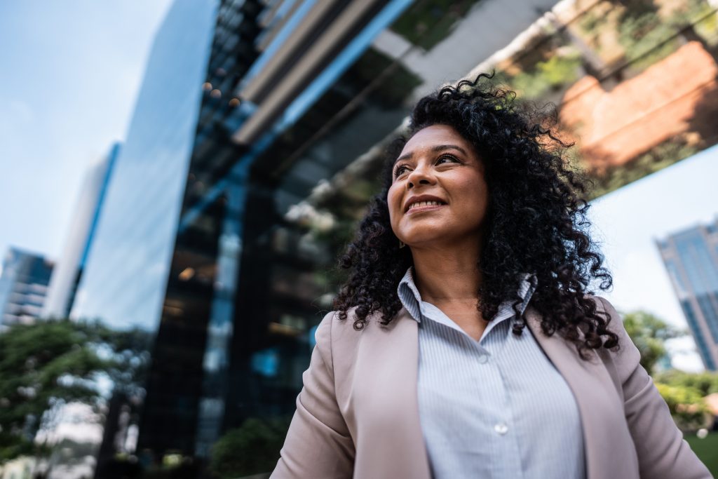 Motivated businesswoman standing outside a city building, contemplating a career change to own a SYNERGY HomeCare franchise.