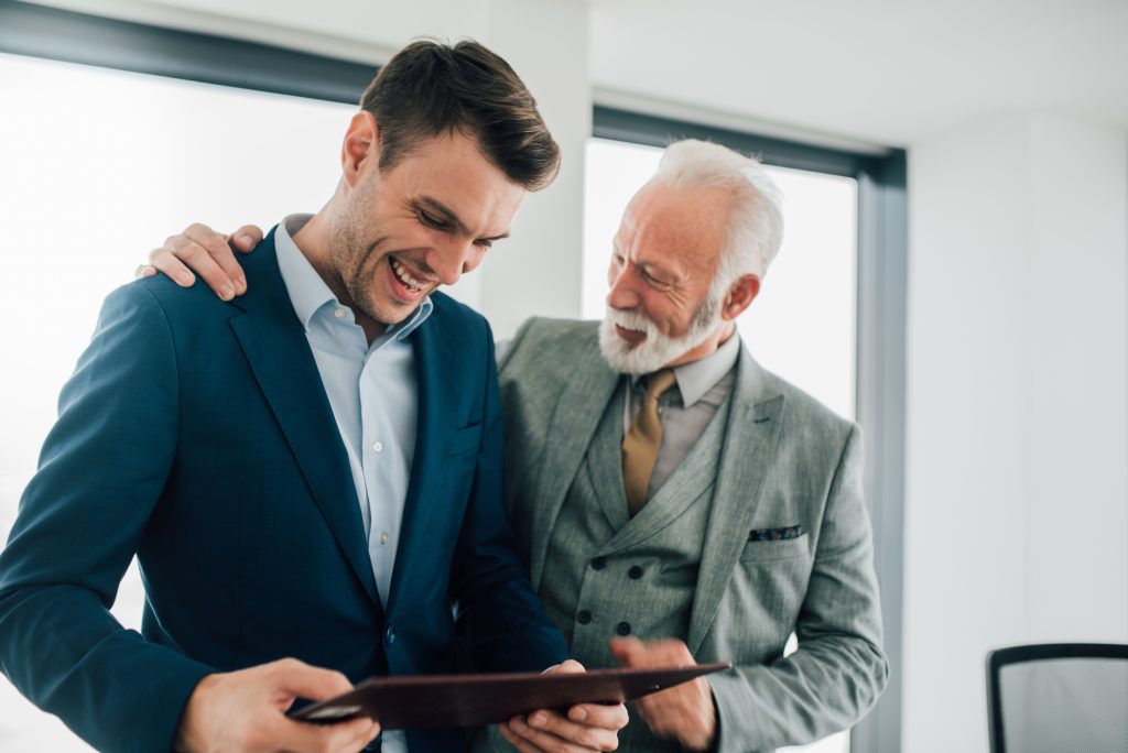 Entrepreneur father and son smiling and standing together in an office as they look at a business document on a clipboard.
