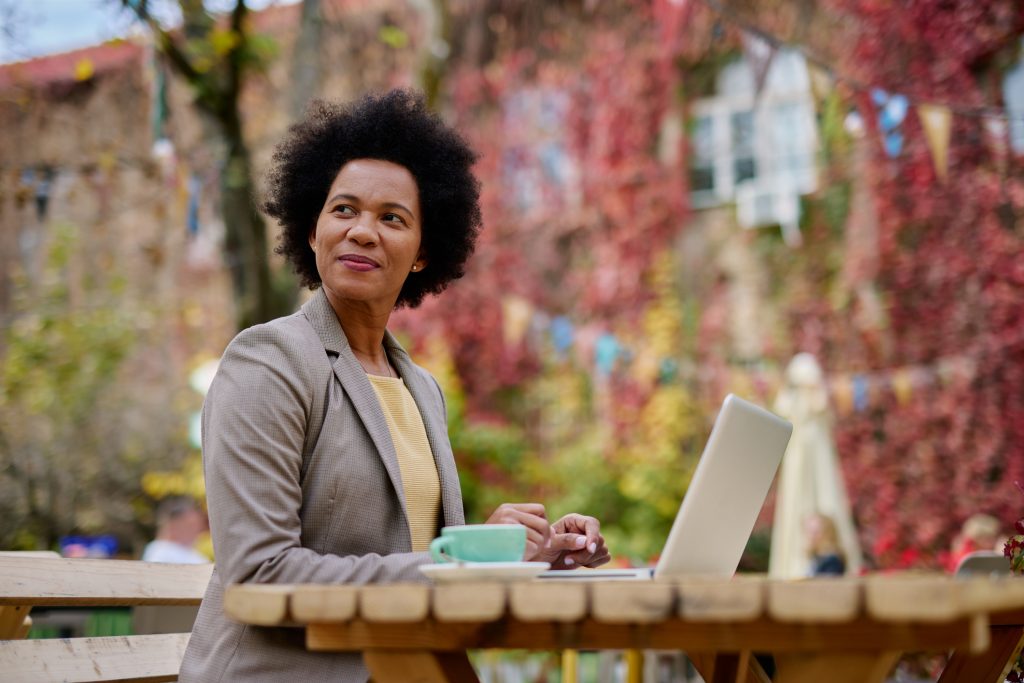 A female entrepreneur dressed in business attire is sitting at an outdoor cafe table with her laptop and a cup of coffee. She looks optimistically into the distance and is surrounded by colorful autumn foliage.