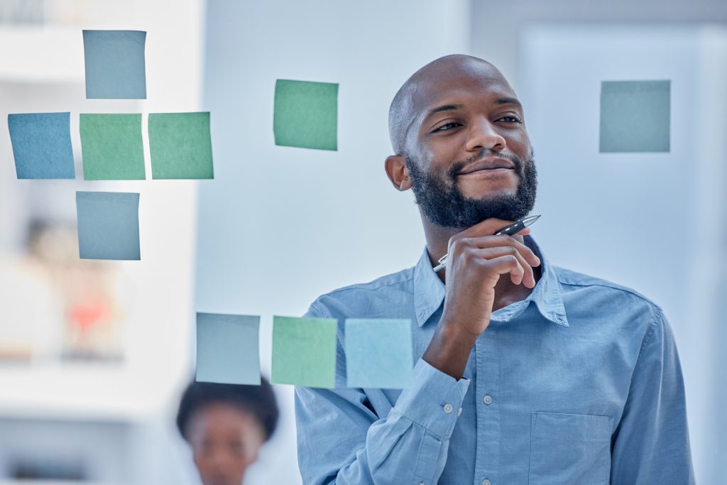 Black male entrepreneur using sticky notes on glass board to organize brainstormed business ideas.