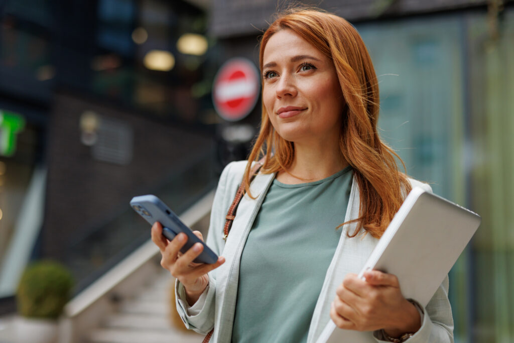 A thoughtful millennial woman entrepreneur holds a smartphone and laptop, standing outside an urban building, looking into the distance with an optimistic smile.