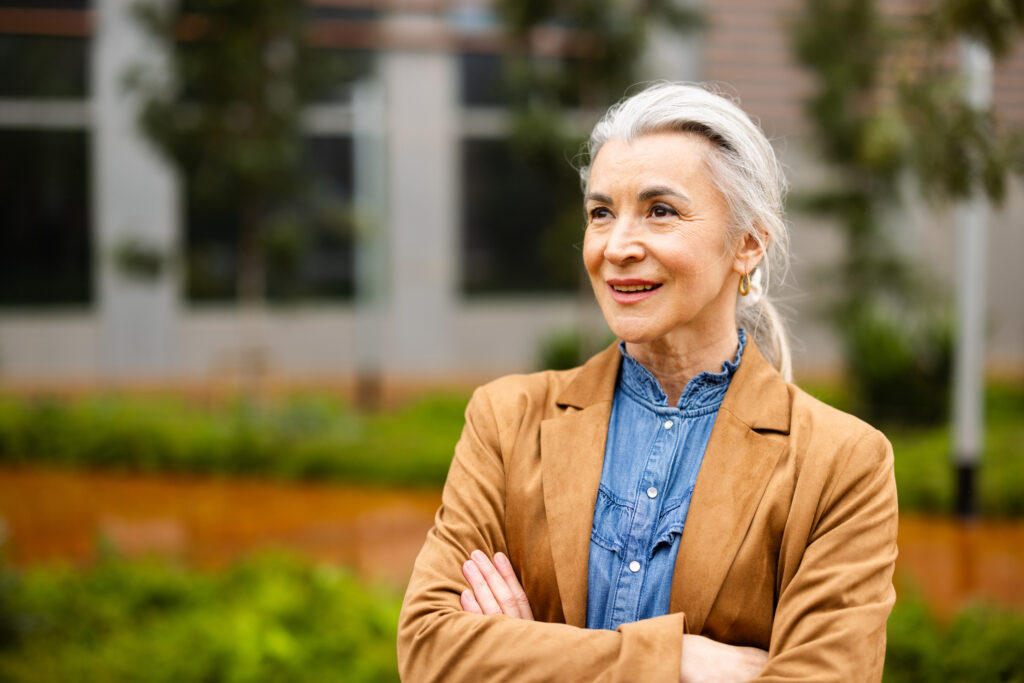 Confident senior adult woman entrepreneur with gray hair smiling with arms crossed looking away outdoors