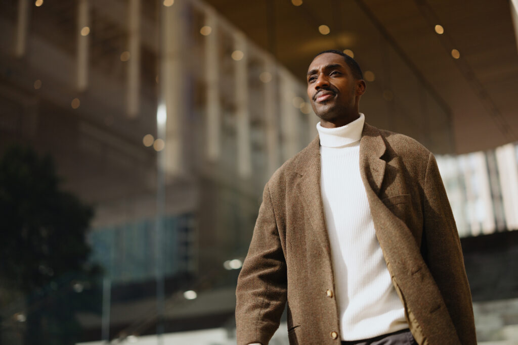 A confident male professional walks through a modern building exterior in soft lighting, while expressing hopefulness and embodying a sense of purpose
