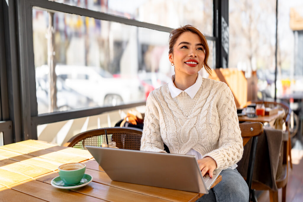 Optimistic, smiling businesswoman looking contemplatively into the distance while using laptop and drinking coffee in outdoor cafe.